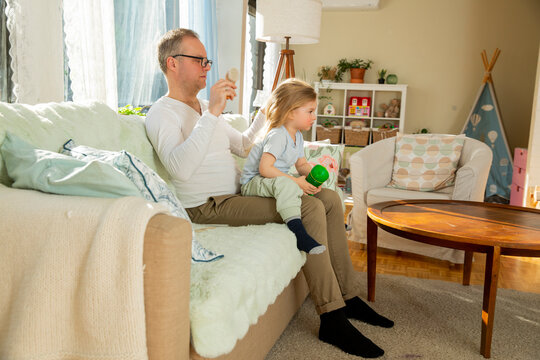 Mature father in glasses brushing long hair of his small child sitting on lap in cozy living room, authentic parenting and analog childhood moments