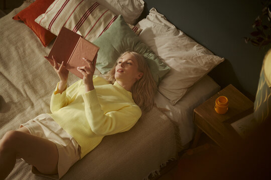 Young Caucasian woman lying on bed and reading book, relaxing at home. Image represents mental wellbeing and self care, suitable for lifestyle and leisure marketing, high angle view