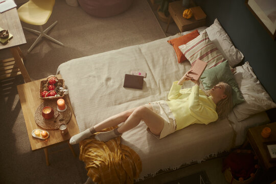 Young Caucasian woman lying on bed and reading book, relaxing in cozy bedroom. High angle view image suitable for mental wellbeing and self-care marketing campaigns