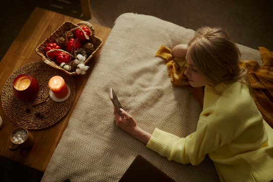 Caucasian young woman lying on bed and using smartphone, browsing social media. Image represents digital life and is suitable for mobile app marketing, high angle shot