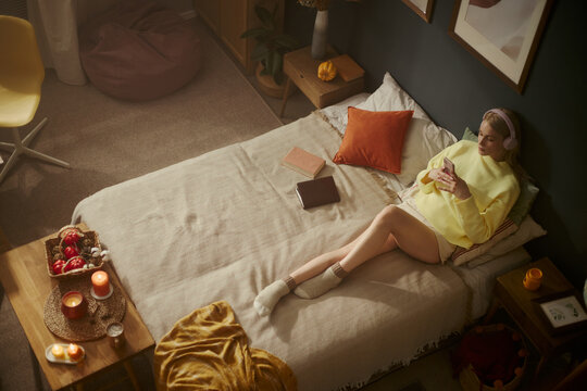 Young Caucasian woman lying on bed wearing headphones and using smartphone. Image represents digital lifestyle and mental wellbeing, suitable for mobile app marketing, high angle shot