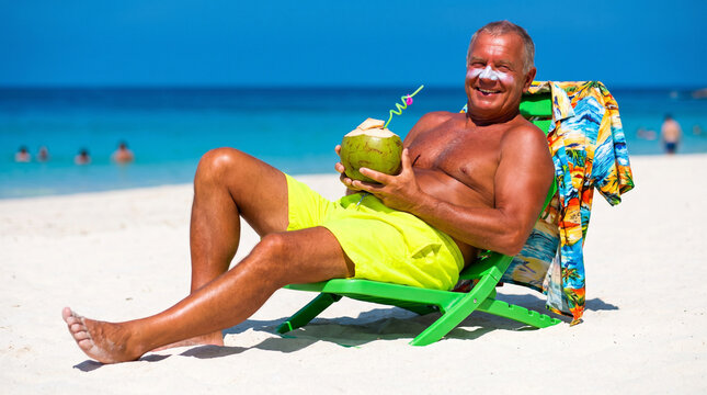 Happy senior man with sunscreen cream on his nose relaxing on a sun lounger at a tropical white sand beach holding a fresh green coconut drink.
