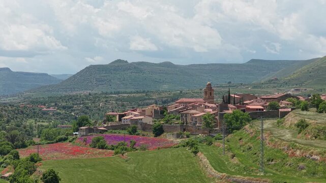 Wide panoramic shot of the historic village of Mirambel in Spain, surrounded by lush green hills and vibrant spring wildflower fields.