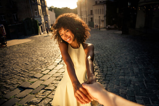 Young woman smiling and holding partner's hand while walking through a historic cobblestone street at sunset, enjoying a romantic travel moment