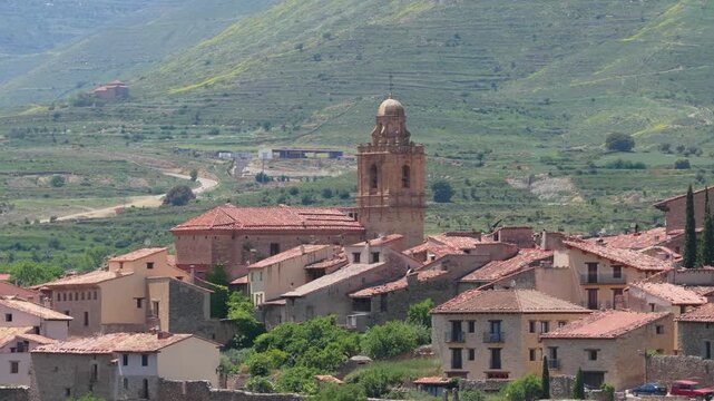 A picturesque view of the historic village of Mirambel in Spain, featuring traditional stone houses with red-tiled roofs clustered around a prominent church tower.