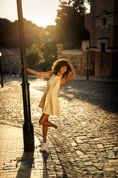 Young woman in yellow dress smiling and posing by a lamppost on a cobblestone street at golden hour, urban summer portrait
