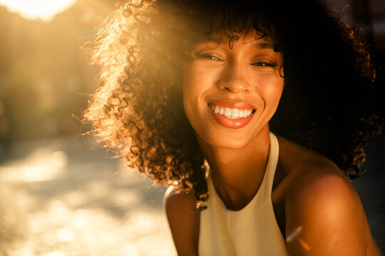 Smiling young woman portrait with curly hair basking in warm golden hour sunlight outdoors, natural beauty and joyful expression with soft lens flare