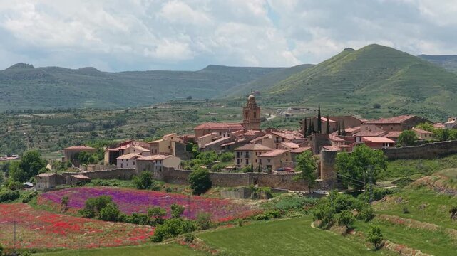 A scenic aerial view of the historic village of Mirambel, Spain, surrounded by colorful wildflower fields and lush green hills.