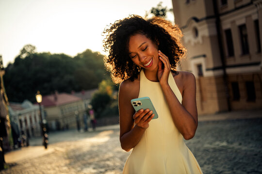 Young smiling woman reading a message on her smartphone while walking on a sunlit cobblestone city street at golden hour enjoying a warm summer evening