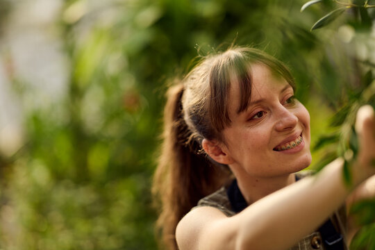Smiling Young Woman Gardening in Greenhouse Reaching for Plants, Natural Outdoor Lifestyle and Growth
