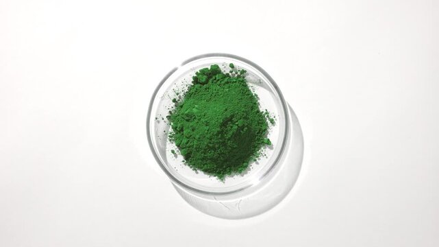 Overhead shot of a pile of green powder held in a glass petri dish on a soft gray backdrop lit with orbiting light and casts moving shadow