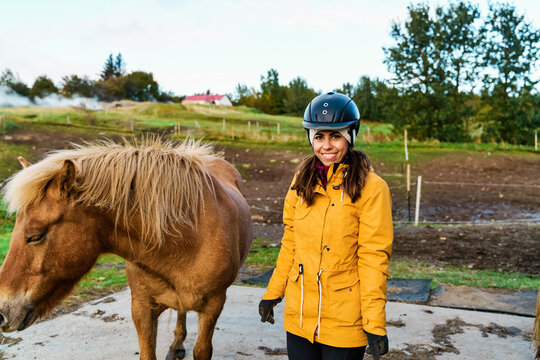 A white woman stands smiling beside a brown horse on a farm