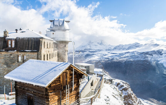 The Gornergrat Observatory sits on a rocky plateau in Zermatt, Switzerland, surrounded by snow covered peaks, including the Matterhorn, under a clear blue sky.