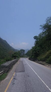 Scenic driving footage of a paved mountain highway winding through lush green forested hills in Nepal. The road passes through a beautiful natural landscape beneath a clear blue sky