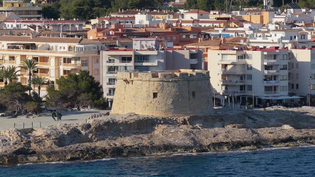 A stone watchtower stands on a rocky coastline in Moraira, Spain, with the Mediterranean Sea in the foreground.