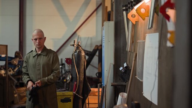 Young adult Caucasian male welder opening locker, putting on protective gloves and safety face shield in workshop. Clip is ideal for industrial safety training and manufacturing workforce videos