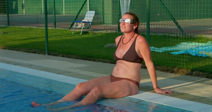 Relaxing poolside, a woman in swimwear enjoys the sun's warmth, her eyes closed in peaceful repose as her feet dangle in the cool water. The scene evokes a sense of summer leisure and relaxation.