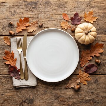 Rustic autumn table setting with white plate, gold cutlery and fall leaves. Thanksgiving dinner mockup on wooden background with pumpkin.
