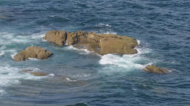 Dynamic footage of ocean waves crashing and swirling around rugged coastal rocks along the shoreline of Baiona, Galicia, Spain.