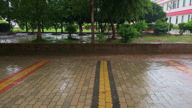 Static shot of raindrops falling onto wet brick pavement and puddles with lush trees and a landscaped park in the background