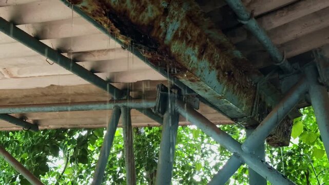 Close up view beneath an old roof showing rusted metal beams pipes and water dripping through the weathered structure