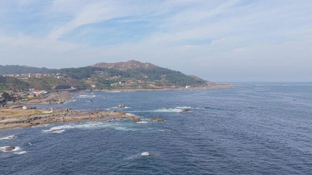 Wide aerial view of the rugged coastline near Baiona, Spain, featuring rocky shores, blue ocean waves, and a scenic hillside with scattered houses. No people are visible in the scene.