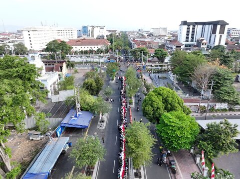 Yogyakarta, Indonesia - 15 August 2026: Aerial view of a long line of people participating in a parade along a street in the city, surrounded by urban buildings and lush trees.