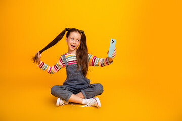 Naklejka na ściany i meble Young girl with pigtails taking a playful selfie on a smartphone while sitting cross-legged against a bright yellow background