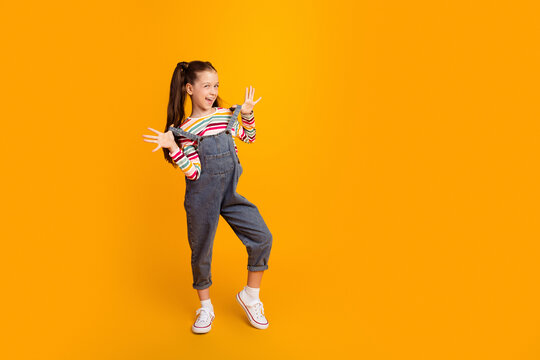 Happy young girl in denim overalls and striped shirt striking playful pose on bright orange background