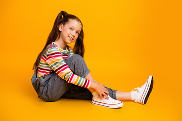 Smiling girl in colorful striped shirt and denim overalls sitting on an orange studio background...