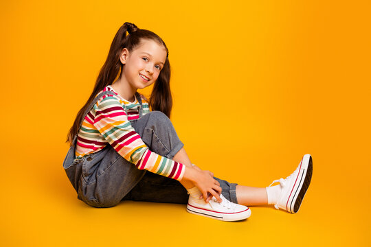Smiling girl in colorful striped shirt and denim overalls sitting on an orange studio background &mdash; casual youthful portrait of a happy child in white sneakers