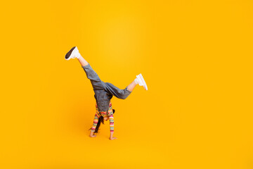 Fototapeta premium Young girl doing a handstand in denim overalls and white sneakers on a bright yellow background, playful energetic childhood movement and joy