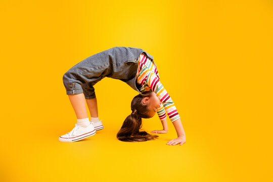 Young girl doing a backbend bridge pose on a bright yellow background showing flexibility, playfulness and active childhood in casual denim overalls and striped shirt