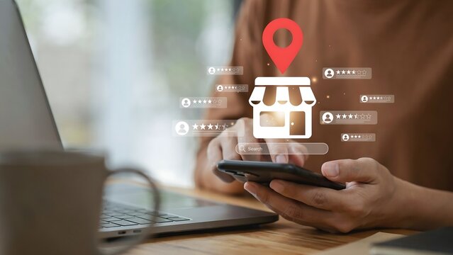 A person uses a smartphone to search for a nearby store, represented by a location pin icon above a shop illustration. The user is in a home or office setting with a laptop and a mug.