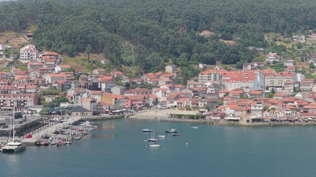 A high-angle aerial shot of the coastal village of Combarro, Spain, showing its picturesque waterfront, marina with docked boats, traditional red-roofed houses, and lush green hills in the background.