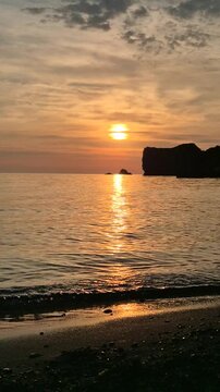 Golden sunset ocean view with rocky cliffs on the horizon at dusk on a sandy beach. Static wide shot.