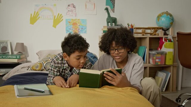 African American teenager and child lying on bed, reading book together in bedroom. Clip is perfect for educational campaigns, tutoring services, and family-oriented marketing