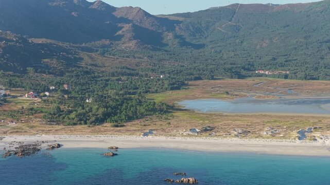 A sweeping aerial shot captures the stunning coastline of Carnota, Spain, featuring the expansive sandy beach, turquoise Atlantic waters, winding lagoon, and lush green mountains in the background.
