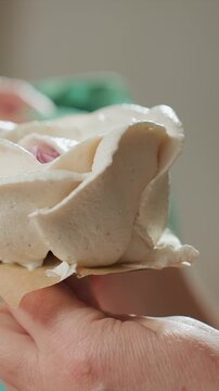 Close up of hand holding white rose shaped pastries with creamy frosting. Focus remains on detailed texture petals. Background shows blurred green objects suggesting another person