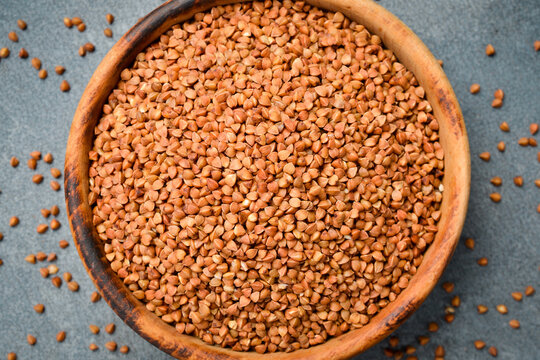 Dry buckwheat cereal is served in a natural timber dish. Healthy culinary ingredient is centered on a grey background. Flat lay perspective of raw food.