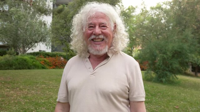Elderly caucasian man with long curly white hair and beard smiling happily outdoors in a green park