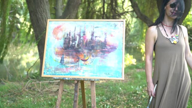 Beautiful woman artist painting on a canvas by the river in a dress