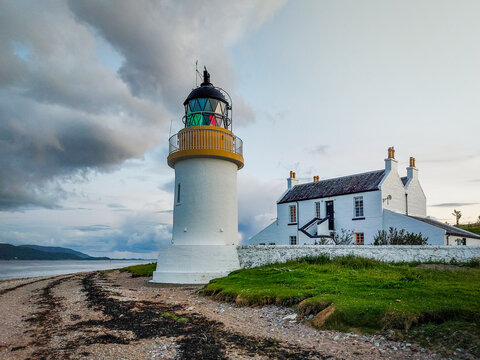 View of Corran Point Lighthouse with its white tower and yellow trim, standing beside a white cottage on the rocky shore under a cloudy sky in Fort William, Scotland, United Kingdom.