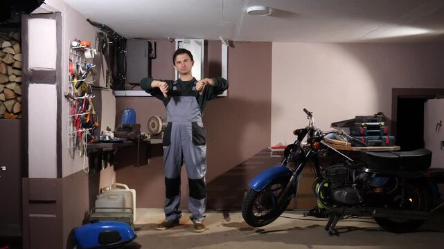 Male mechanic wearing overalls standing in a cluttered garage or workshop, giving two thumbs down, expressing failure, dissatisfaction or negative feedback while working on a motorcycle