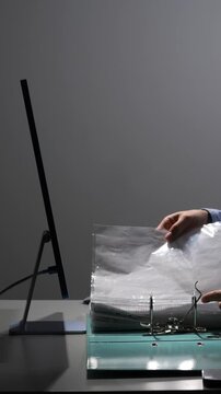 Vertical video person's hands meticulously organizing paper documents into a ring binder at a workstation, with a computer monitor visible, symbolizing administrative tasks and workflow