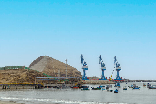 View of Puerto Chancay with large blue industrial cranes, a pier, and small fishing boats in the harbor in Chancay, Lima Region, Peru.