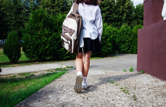 Schoolgirl walks on a pathway leading to school. She carries a backpack and wears a school uniform. It is daytime