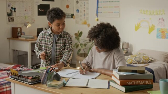 Black teenage boy writing in notebook at desk while younger African American brother stands nearby eating apple and discussing homework. Clip ideal for e-learning and tutoring ads