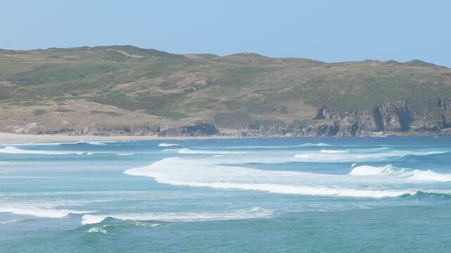 Wide shot of Praia do Rostro beach in Spain, showing turquoise waves rolling onto the sandy shore with rugged green hills in the background.