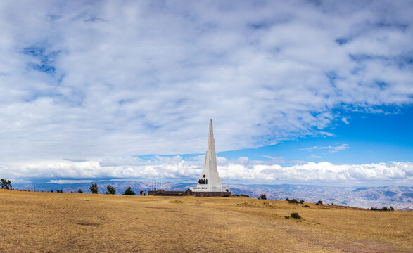 View of the Pampa De La Quinua monument standing in a vast, dry, golden-brown field under a bright blue sky with scattered white clouds in Quinua, Ayacucho, Peru.